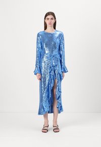 Duga plava haljina od šljokica s dugim rukavima, nabranim rubom i prednjim prorezom, vezana u struku odgovarajućom trakicom. Ravne crne sandale.