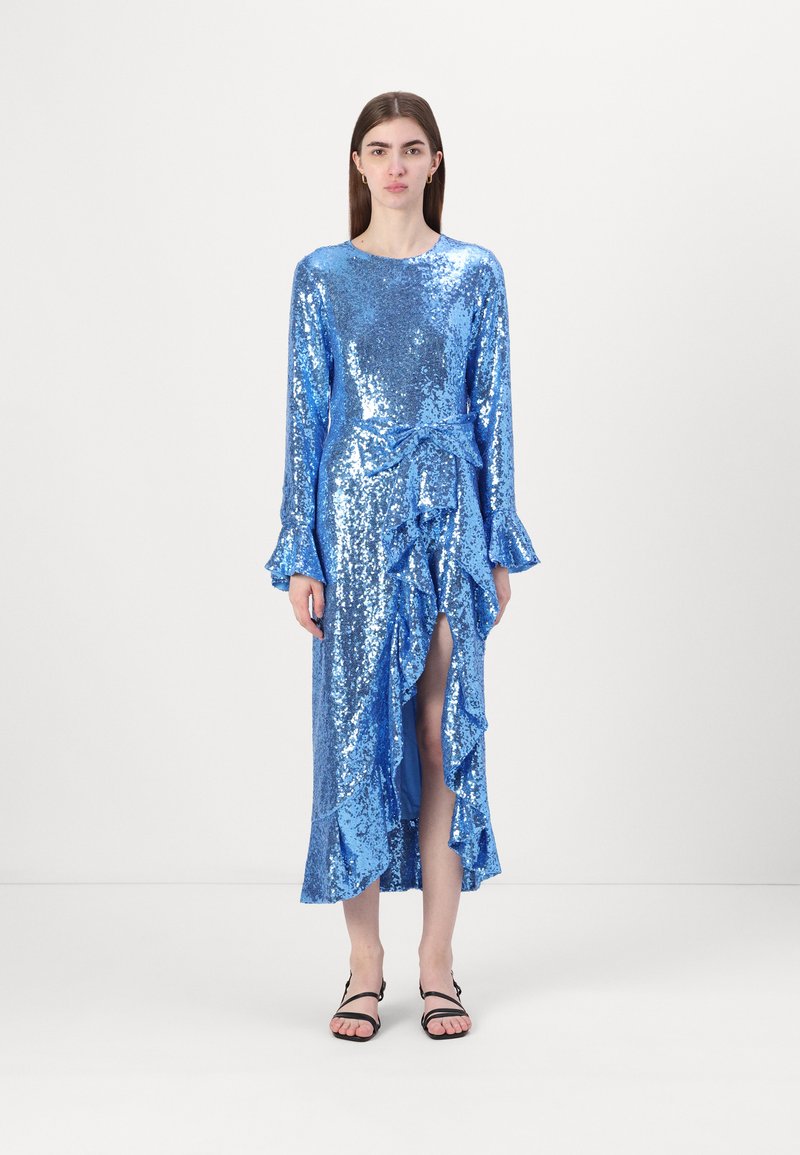 Duga plava haljina od šljokica s dugim rukavima, nabranim rubom i prednjim prorezom, vezana u struku odgovarajućom trakicom. Ravne crne sandale.