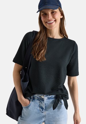 Kvinde iført en sort tekstureret kortærmet top bundet om taljen, lyseblå jeans, blå kasket og bærer en sort skuldertaske.