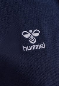 Granatowy materiał z haftowanym białym logo, które przedstawia symetryczny wzór przypominający pszczołę z skrzydłami oraz słowo "hummel" poniżej.