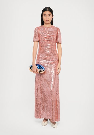 Vrouw in een vloerlang, roze, paillettenkleed met een metalen ovale clutch, staand tegen een witte achtergrond met een neutrale uitdrukking.