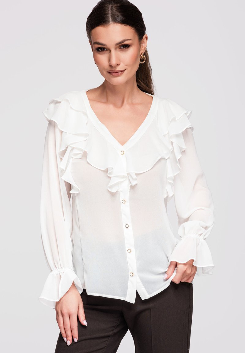 Femme aux longs cheveux foncés porte un chemisier blanc à volants avec des boutons dorés et un pantalon noir, posant devant un fond clair uni.