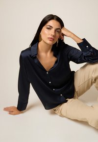 Navyblaue Satinbluse mit Kragen, Vorderknöpfen und langen Ärmeln, kombiniert mit beigen Hosen. Das Model posiert mit einer Hand im Gesicht.