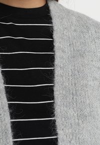 Cárdigan de punto gris claro con un acabado texturizado, usado sobre una camiseta negra con rayas horizontales blancas.
