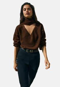 Ruskea, teksturoitu neulepaita, jossa on syvä V-pääntie ja huivielementti. Päällä tummien, istuvien housujen kanssa, jotka on kiinnitetty mustalla vyöllä, jossa on kullanvärinen solki.