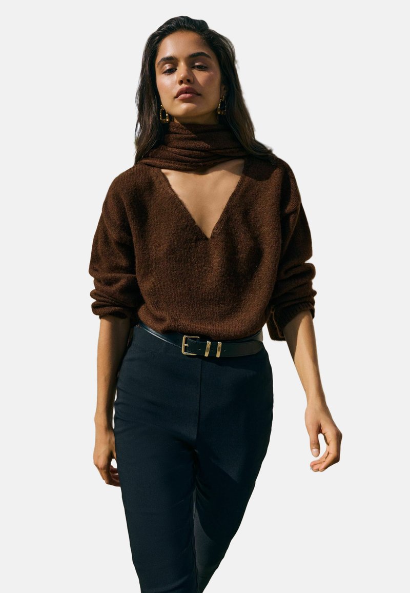 Ruskea, teksturoitu neulepaita, jossa on syvä V-pääntie ja huivielementti. Päällä tummien, istuvien housujen kanssa, jotka on kiinnitetty mustalla vyöllä, jossa on kullanvärinen solki.