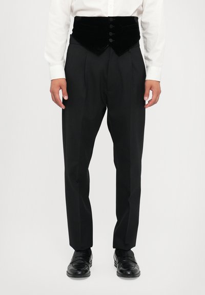 Un pantalon de smoking noir avec un plastron en velours taille haute comportant six boutons, associé à une chemise blanche et des chaussures noires.