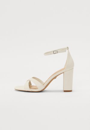 Cream-colored heeled sandal with a square toe, crisscross straps, ankle strap with buckle, and a chunky heel. Smooth leather texture.