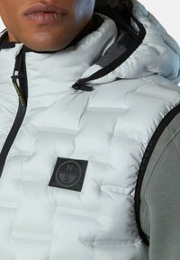 Weißer Daunenweste mit hohem Kragen, ausgestattet mit einem schwarzen Reißverschluss und einem Logo-Patch. Die Außenseite ist gesteppt und hat eine glatte, glänzende Oberfläche.