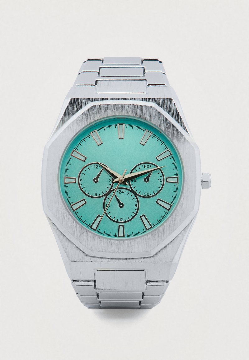 Montre en acier inoxydable argentée avec un boîtier octogonal, un cadran turquoise, trois sous-cadrans et un bracelet en métal brossé.
