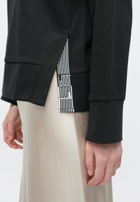 Μαύρο μακρυμάνικο τοπ με πλευρική σχισμή που διαθέτει μαύρες και λευκές ριγέ λεπτομέρειες. Μαλακό ύφασμα με άνετη εφαρμογή και μινιμαλιστικό σχέδιο.