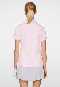 Polo skjorte i lys pink med krave og korte ærmer, sat sammen med en grå nederdel. Stoffet ser glat ud uden synlige mønstre.