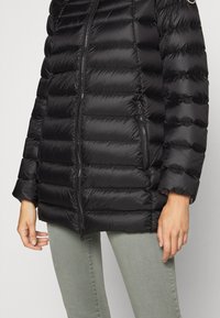 Černá péřová bunda s prošívaným designem, zapínáním na zip a bočními kapsami. Měkká, lesklá textura, s kulatým logem na rameni.