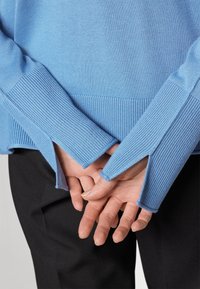 Die Hände hinter dem Rücken verschränkt, getragen wird ein hellblaues, geripptes Sweatshirt mit verlängerten, geschlitzen Bündchen und schwarze Hosen.