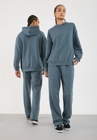 Mėlyngray hoodie ir atitinkamos sportinės kelnės iš minkštos medvilnės mišinio; laisvas kirpimas, susiraukšlėti rankogaliai ir juosmuo; minimalizmo dizainas be raštų.