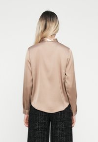 Camicia in satin a maniche lunghe color beige chiaro con colletto lucido. Tessuto morbido, orlo arrotondato e polsini con bottoni. Abbinata a pantaloni neri a trama.