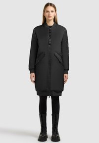 Cappotto imbottito nero con zip frontale, grandi tasche e polsini a costine. Presenta un'etichetta con il logo sulla zip e un design lungo fino al ginocchio.