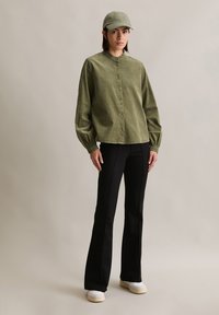 Chemise boutonnée en velours côtelé vert olive avec un col montant, assortie à un pantalon évasé noir et une casquette assortie, arborant un logo brodé.