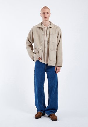 Giacca di cotone beige con bottoni, grande tasca frontale. Camicia leggera sotto, jeans a gamba larga blu e scarpe marroni. Design semplice, colori neutri.