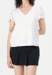 Haut en dentelle blanche avec un col en V et des manches courtes à volants, orné de motifs floraux ; associé à une mini-jupe plissée noire.