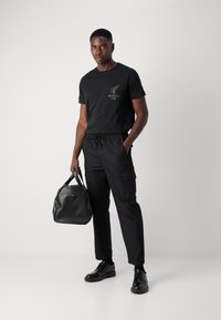 Black T-shirt with green logo, black cargo pants, black dress shoes, and a black tote bag with handles. Smooth fabric and sleek design.