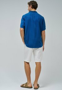 Camisa azul de manga corta hecha de tela ligera, combinada con pantalones cortos blancos. La camisa presenta un clásico cuello y un diseño de yugo trasero.