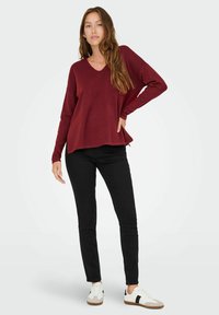 Maglione borgogna a maniche lunghe con scollo a V e una vestibilità morbida, abbinato a jeans skinny neri e sneaker bianche con dettagli neri.