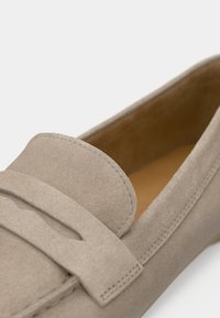 Μπεζ μοκασίνι από σουέντ με επίπεδη σόλα, διακοσμημένο με ραμμένο σχέδιο και διακοσμητικό λουράκι πάνω από το πέλμα. Το εσωτερικό είναι από δέρμα ταμπά.