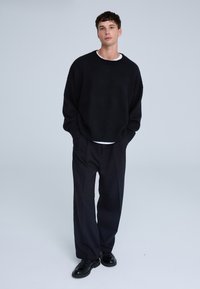 Maglione nero oversize con polsini e orlo a coste, indossato sopra una camicia bianca. Abbinato a pantaloni neri e scarpe nere lucide.