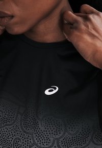 Κοντινό πλάνο ενός ατόμου που προσαρμόζει το γιακά ενός μαύρου αθλητικού μπλουζιού με λευκό λογότυπο Asics και πουά σχέδιο στο ύφασμα.