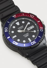 Montre à quartz Casio avec bracelet en caoutchouc noir, boîtier rond, cadran noir, aiguilles argentées et lunette rotative rouge-bleue. Résistante à l'eau jusqu'à 100m.