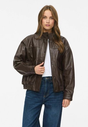 Modèle portant une veste en cuir marron foncé sur un haut blanc et un jean bleu, tenant le devant de la veste devant un fond blanc uni.