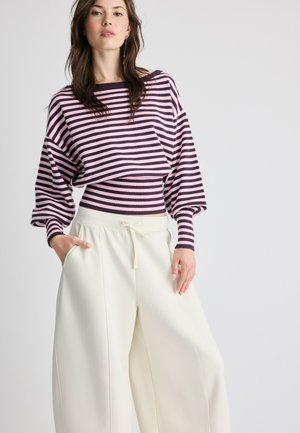 Jeune femme portant un pull rayé rose et noir avec des manches bouffantes et un pantalon blanc ample taille haute avec un cordon.
