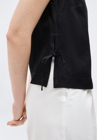 Πλάγια όψη ενός ατόμου που φοράει μαύρο αμάνικο τοπ με δεμένη πλαϊνή σχισμή και λευκό παντελόνι μπροστά σε απλό φόντο.