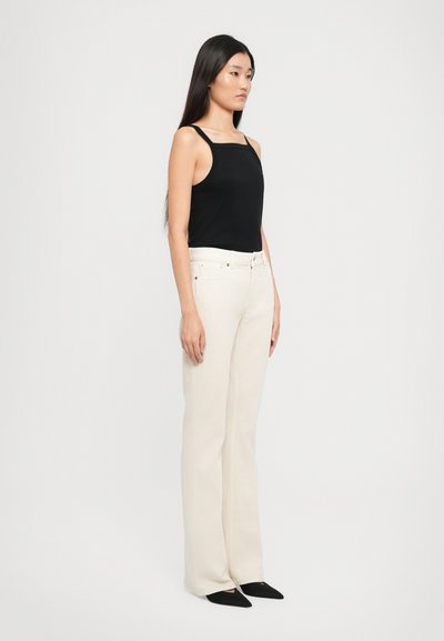 Mujer de pie de lado, vistiendo una blusa negra sin mangas, pantalones anchos de tiro alto color blanco y zapatos negros de punta en un fondo blanco.
