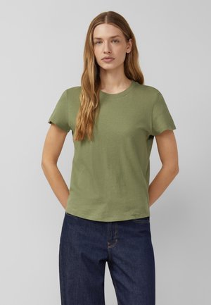 Kobieta stoi ze skrzyżowanymi za plecami rękami, ubrana w oliwkowo zieloną koszulkę z krótkim rękawem i ciemnoniebieskie dżinsy na jednolitym szarym tle.