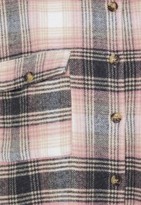 Plaid flannel shirt in pink, gray, and beige. Features buttons and a chest pocket with contrasting patterns and textures.