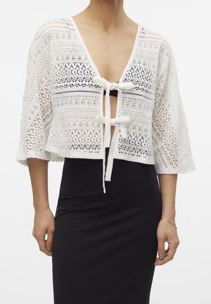 Femme portant un cardigan blanc en crochet à nouer devant avec des manches trois-quarts, par-dessus une jupe ou une robe noire ajustée.