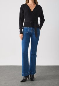 Schwarzer Wickelpullover mit langen Ärmeln und gebundenem Gürtel, kombiniert mit dunkelblauen, ausgestellten Jeans mit ausgefransten Säumen und spitzen schwarzen Stiefeletten.