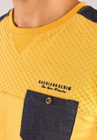 Maglione giallo con superficie testurizzata, tasca in denim scuro e accenti a contrasto sulle spalle. Presenta il logo del brand ricamato in blu navy.