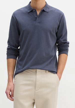 Homme portant un polo bleu foncé à manches longues avec deux boutons et un pantalon beige, une main dans la poche, sur un fond uni.