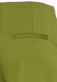 Gros plan sur un vêtement en tissu vert avec une poche fendue horizontale et des coutures visibles sur une surface lisse.