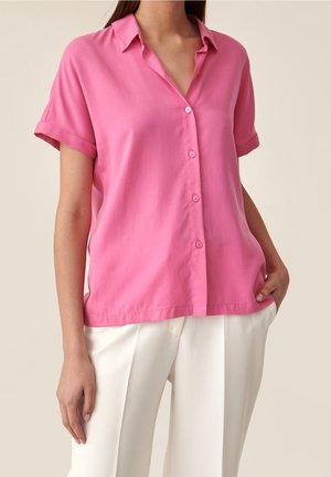 Camicetta a maniche corte con colletto in tessuto rosa brillante, con bottoni frontali e vestibilità comoda, abbinata a pantaloni bianchi.