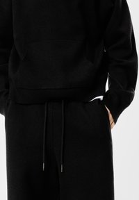 Fekete pamut pulóver kenguru zsebbel, bordás aljjal és mandzsettával, fekete tréningnadrággal párosítva, amely húzózsinórral és oldalzsebekkel rendelkezik.