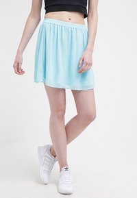 Donna con una gonna azzurro chiaro, top nero alla vita e scarpe bianche Adidas, in piedi con le gambe incrociate.