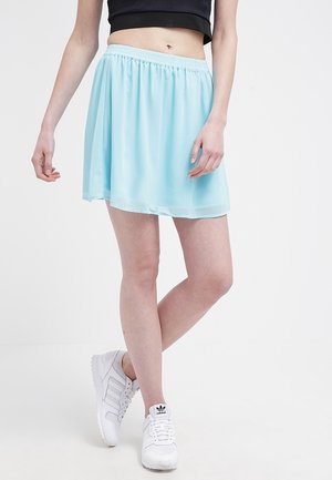 Mujer vestida con una falda azul clara, un top corto negro y zapatillas Adidas blancas, de pie con las piernas cruzadas.