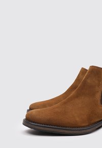 Botas de tobillo de ante marrón con una textura suave, paneles laterales elásticos y suelas oscuras contrastantes. Diseño clásico y minimalista con forma de punta redondeada.