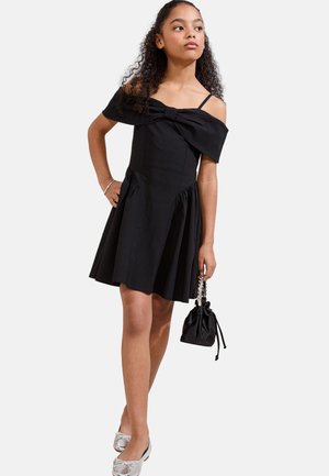 Chica con vestido negro con los hombros descubiertos y detalle de lazo, sosteniendo un pequeño bolso negro, llevando zapatos planos plateados, de pie con la mano en la cadera.