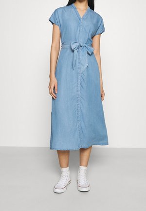 Femme portant une robe midi en jean bleu clair avec manches courtes, une ceinture nouée à la taille et des baskets blanches montantes.