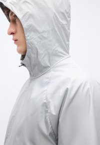 Ανοιχτό γκρι μπουφάν με κουκούλα, με λείο, γυαλιστερό ύφασμα και φερμουάρ μπροστά. Διαθέτει μινιμαλιστικό σχέδιο και διακριτικό λογότυπο στο στήθος.